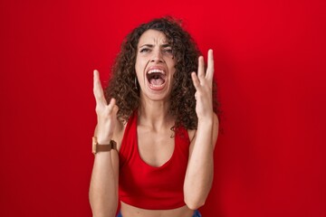 Hispanic woman with curly hair standing over red background crazy and mad shouting and yelling with aggressive expression and arms raised. frustration concept.