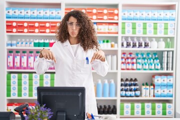 Hispanic woman with curly hair working at pharmacy drugstore pointing down looking sad and upset,...