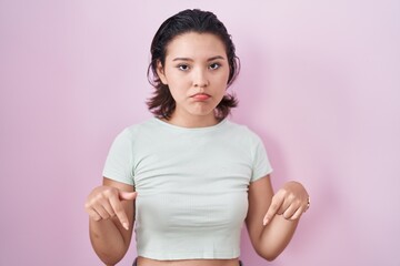 Hispanic young woman standing over pink background pointing down looking sad and upset, indicating...
