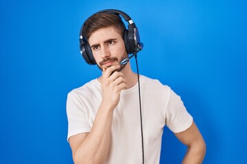 Hispanic man with beard listening to music wearing headphones with hand on chin thinking about question, pensive expression. smiling with thoughtful face. doubt concept.