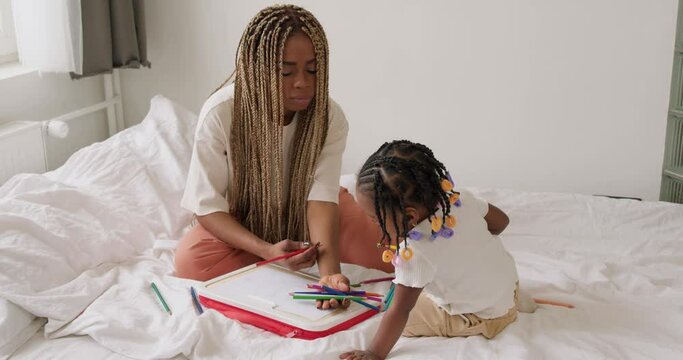 beautiful mother and daughter happily playing in bed together. Mother teaches teaches children to draw. mom giving colored pencil to kid free time spare time lifestyle hobby slow motion