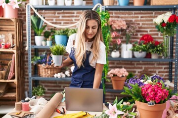 Young blonde woman florist using laptop holding plant at florist