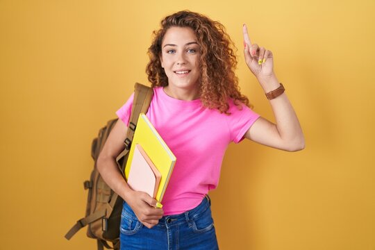 Young caucasian woman wearing student backpack and holding books smiling amazed and surprised and pointing up with fingers and raised arms.