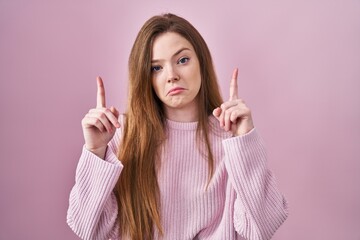 Young caucasian woman standing over pink background pointing up looking sad and upset, indicating...