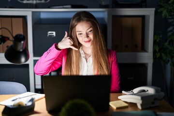 Young caucasian woman working at the office at night smiling doing phone gesture with hand and fingers like talking on the telephone. communicating concepts.