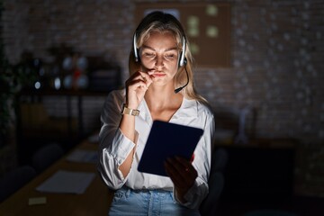 Young blonde woman working at the office at night mouth and lips shut as zip with fingers. secret...