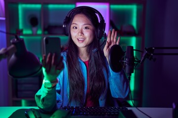 Young asian woman playing video games with smartphone waiving saying hello happy and smiling, friendly welcome gesture