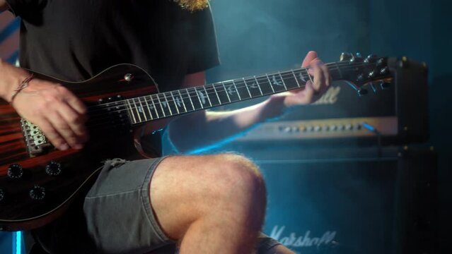 The young musician's hands play electro guitar in a dark studio with a light at the back and a moving camera.