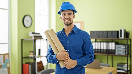 Young hispanic man architect wearing hardhat holding blueprints at office