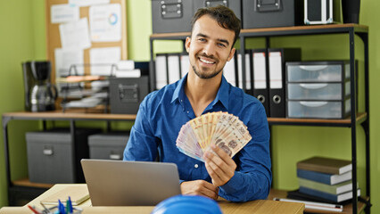 Young hispanic man architect using laptop holding mexican pesos at office