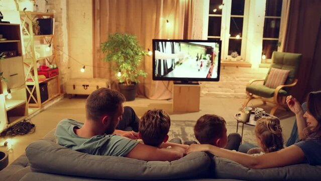 Back view of parents and children spending evening together, sitting on couch in living room and watching tv, eating snacks. Concept of family, leisure time, relaxation, childhood and parenthood