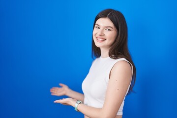 Young caucasian woman standing over blue background inviting to enter smiling natural with open hand
