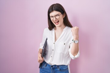 Young caucasian woman holding laptop very happy and excited doing winner gesture with arms raised,...