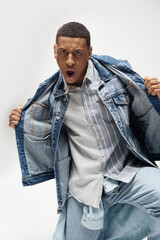 handsome emotional african american man in denim outfit posing on white backdrop, fashion concept