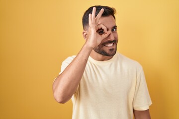 Fototapeta na wymiar Handsome hispanic man standing over yellow background doing ok gesture with hand smiling, eye looking through fingers with happy face.