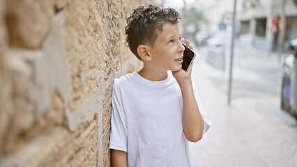 Adorable blond boy engrossed in an important talk on his smartphone, leaning on a wall, street-side...