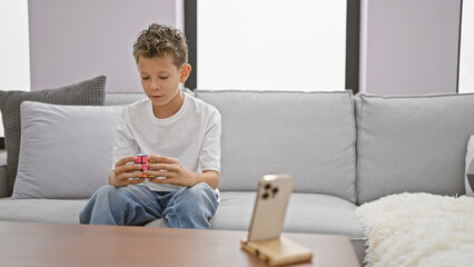 Adorable blond boy sitting on sofa, confidently solving rubik cube puzzle, recording indoor home...