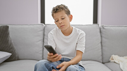 Adorable blond kid wearing a bored expression, comfortably hustling on the sofa at home, seeming...