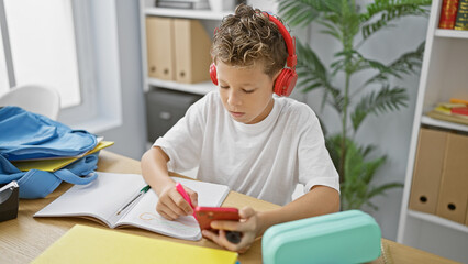 Adorable blond boy student, engrossed in listening to music on his smartphone, diligently drawing...