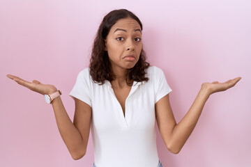 Young hispanic woman wearing casual white t shirt clueless and confused expression with arms and...