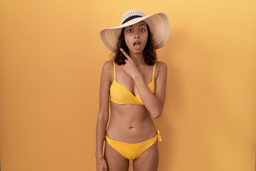 Young hispanic woman wearing bikini and summer hat surprised pointing with finger to the side, open mouth amazed expression.