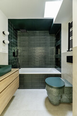 Beautiful bathroom with a modern design with dark green tiling, a curious toilet, hanging wooden furniture, a skylight on the ceiling and black taps