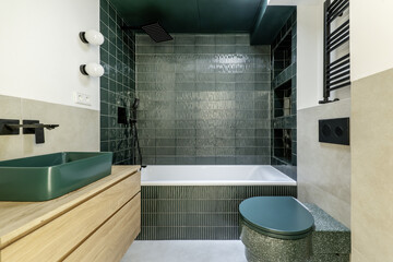 Bathroom of a modern house with white bathtub and glossy dark green tiles, green designer toilet, black towel rack, green porcelain sink on an oak wooden cabinet with drawers