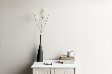 A small white wooden cabinet with a few stacked books, a white mug and a gray vase with artificial branches with subtle white leaves
