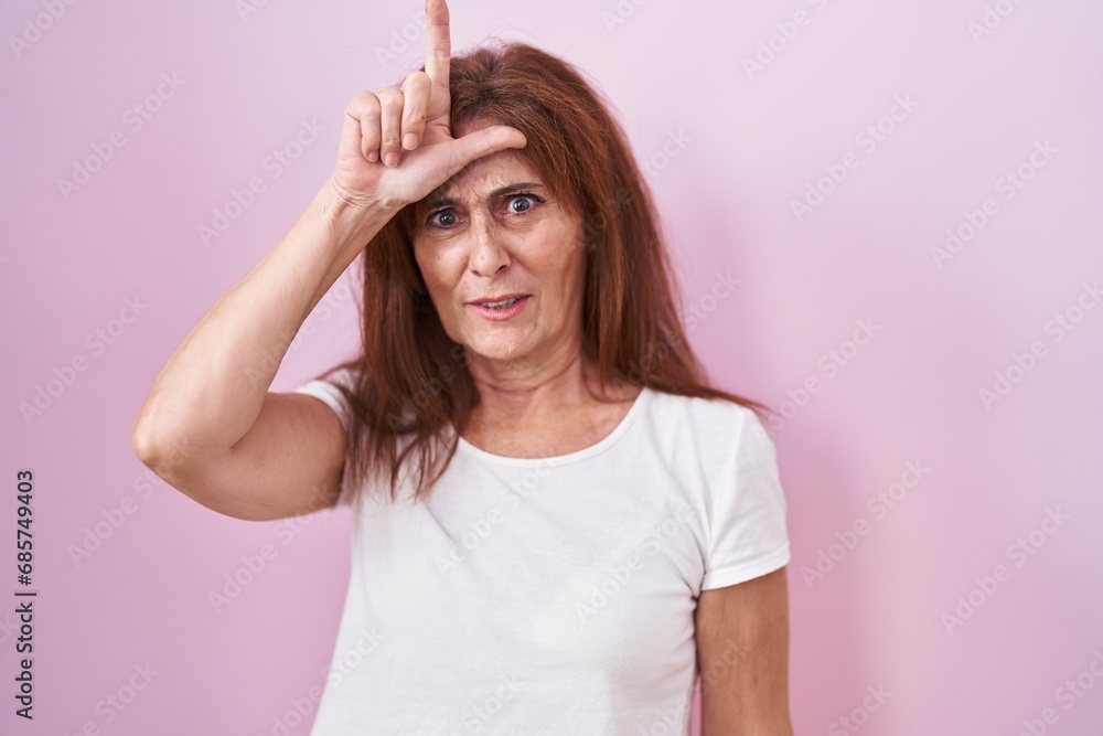 Sticker Middle age woman standing over pink background making fun of people with fingers on forehead doing loser gesture mocking and insulting.