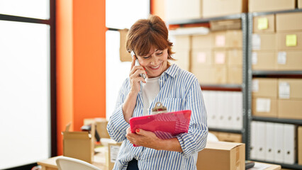 Middle age woman ecommerce business worker reading document talking on smartphone at office