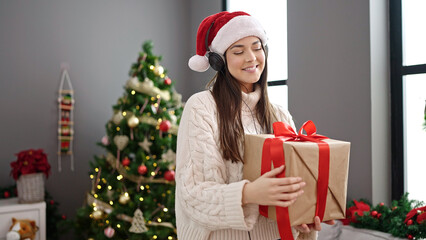 Young beautiful hispanic woman standing by christmas tree holding gift dancing at home