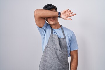 Hispanic young man wearing apron over white background covering eyes with arm, looking serious and...