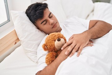Young hispanic man hugging teddy bear lying on bed sleeping at bedroom