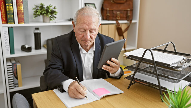 Serious senior businessman, an elegant man with grey hair, diligently working in a relaxed indoor office room, taking notes on his touchpad while sitting at his desk.