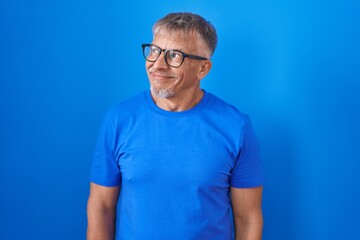 Hispanic man with grey hair standing over blue background smiling looking to the side and staring away thinking.