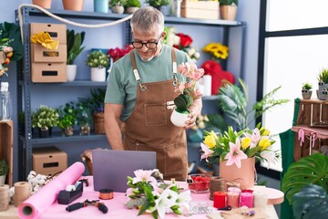 Middle age grey-haired man florist using laptop holding plant at flower shop