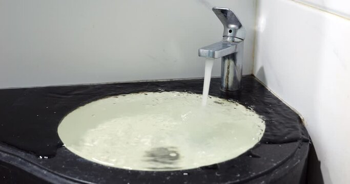 A clog in the bathroom sink is causing the basin to fill up rapidly, leading to an overflow of water onto the bathroom countertop. Old and dirty look of the faucet and room