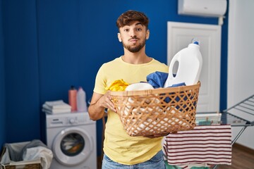 Arab man with beard holding laundry basket and detergent bottle puffing cheeks with funny face. mouth inflated with air, catching air. - obrazy, fototapety, plakaty