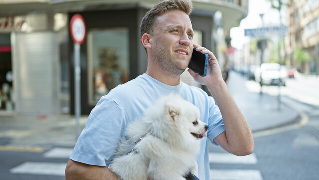 Joyful young caucasian man having a lively talk on his phone while standing on a city street, sharing fun moments with his playful puppy.