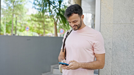 Young hispanic man using smartphone wearing backpack at street