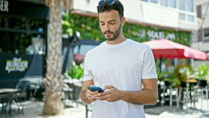 Young hispanic man using smartphone with serious expression at coffee shop terrace