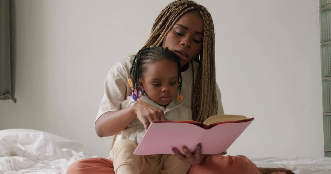 caring african mother reading a book to little daughter on bed at home . black girl sitting with mom study at home in bedroom together quarantine Home School, slow motion education,mom shows pictures