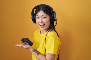 Chinese young woman playing video game holding controller pointing aside with hands open palms showing copy space, presenting advertisement smiling excited happy