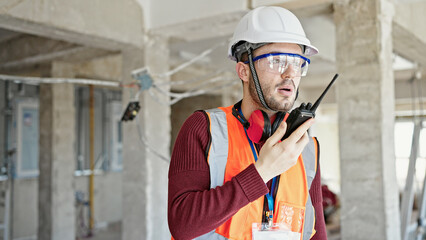 Young hispanic man builder talking on walkie talkie at construction site