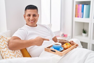 Young hispanic man eating breakfast in the bed smiling happy pointing with hand and finger