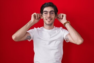 Young hispanic man standing over red background smiling pulling ears with fingers, funny gesture....