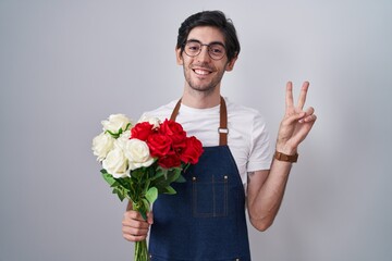 Young hispanic man holding bouquet of white and red roses smiling looking to the camera showing fingers doing victory sign. number two.