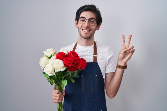 Young hispanic man holding bouquet of white and red roses smiling looking to the camera showing fingers doing victory sign. number two.