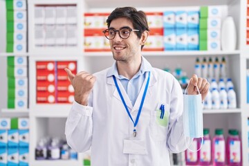 Young hispanic man working at pharmacy drugstore holding safety mask pointing thumb up to the side smiling happy with open mouth