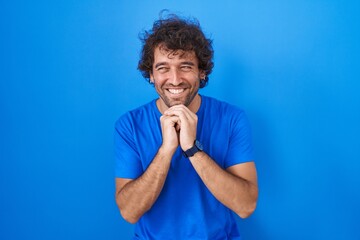 Hispanic young man standing over blue background laughing nervous and excited with hands on chin...